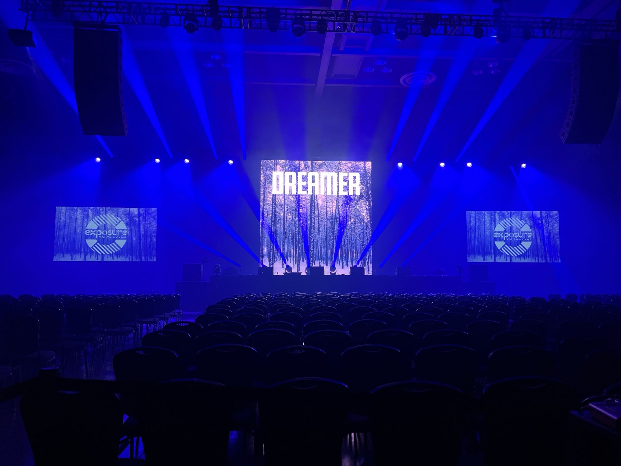 Concert audience facing a stage lit in blue, with large LED screens either side
