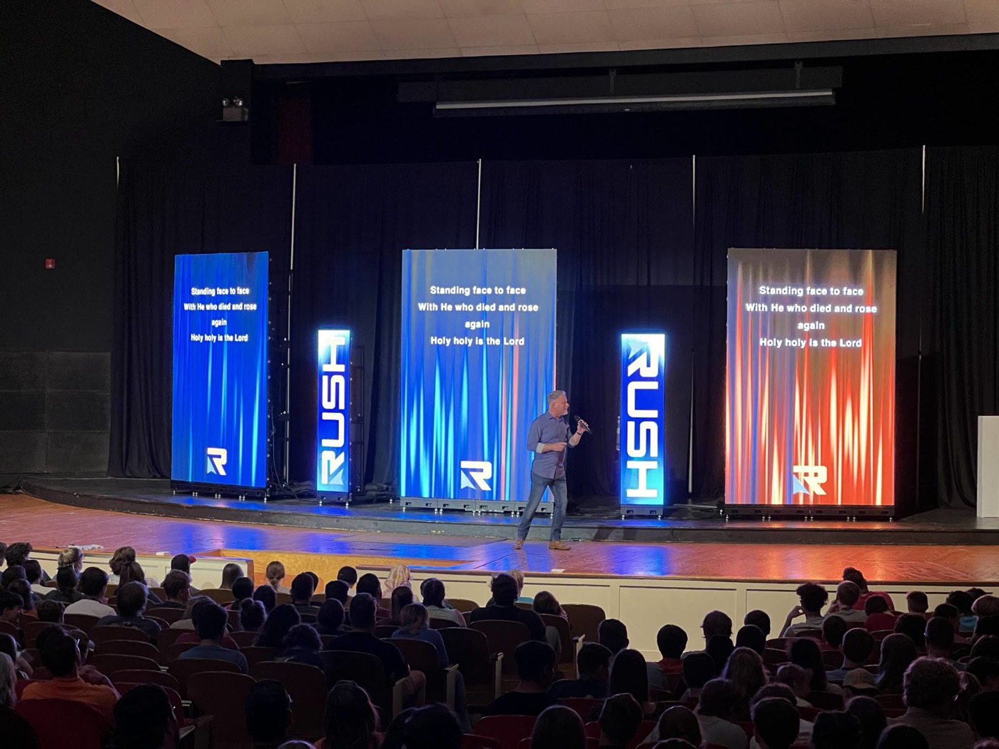 Presenter on stage between three projection screens in front of an audience